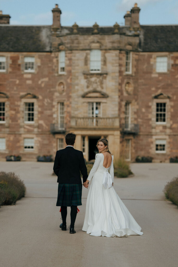 The bride and groom, outside of a large country house. The bride is turning and looking towards the camera.