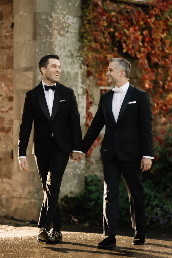 Two men, in black suits, holding hands and smiling at eachother
