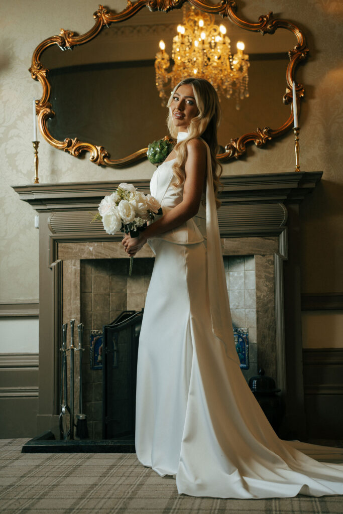 A bride, in her wedding dress, holding a bouquet of flowers in front of a grand fireplace